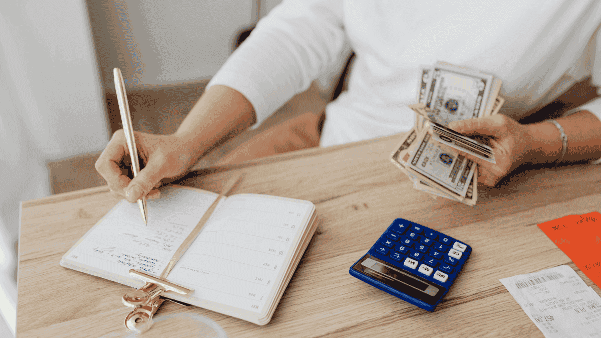 A person writing expenses in a notebook while holding cash, with a calculator nearby
