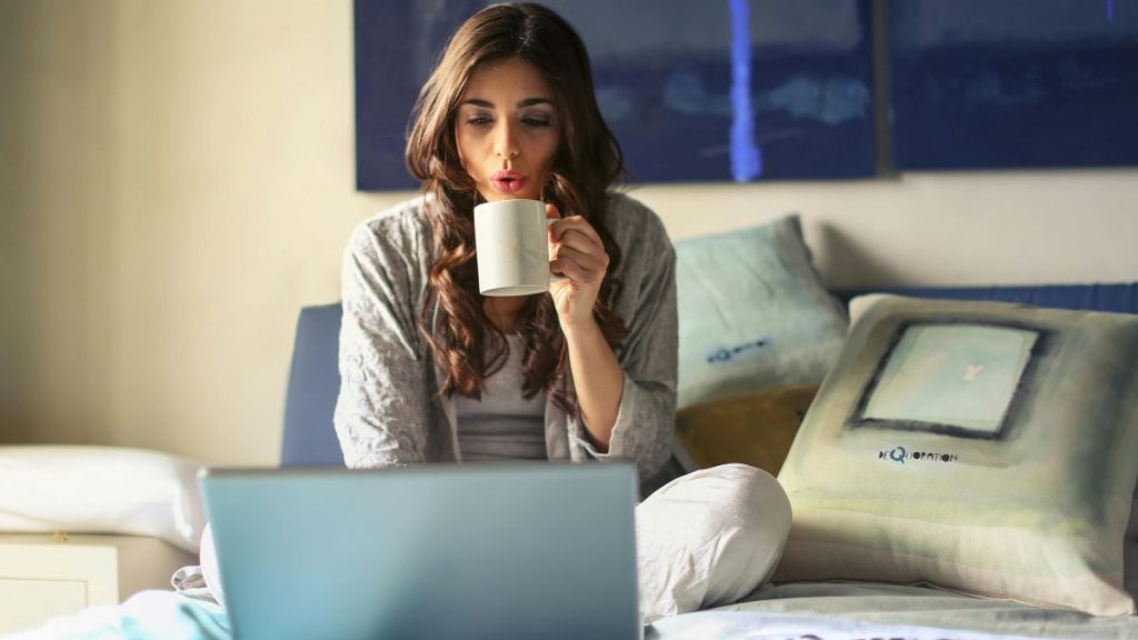 Woman sipping coffee while working on a laptop in bed, symbolizing convenience and modern lifestyle habits.