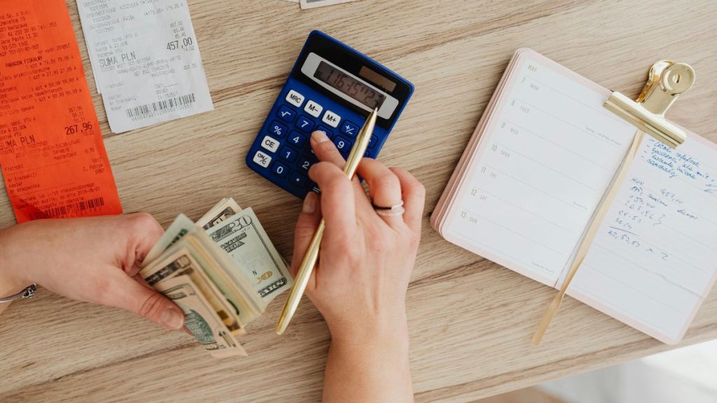 Hands holding cash and using a calculator with a budget planner, showcasing a practical approach to managing debt and personal finances.